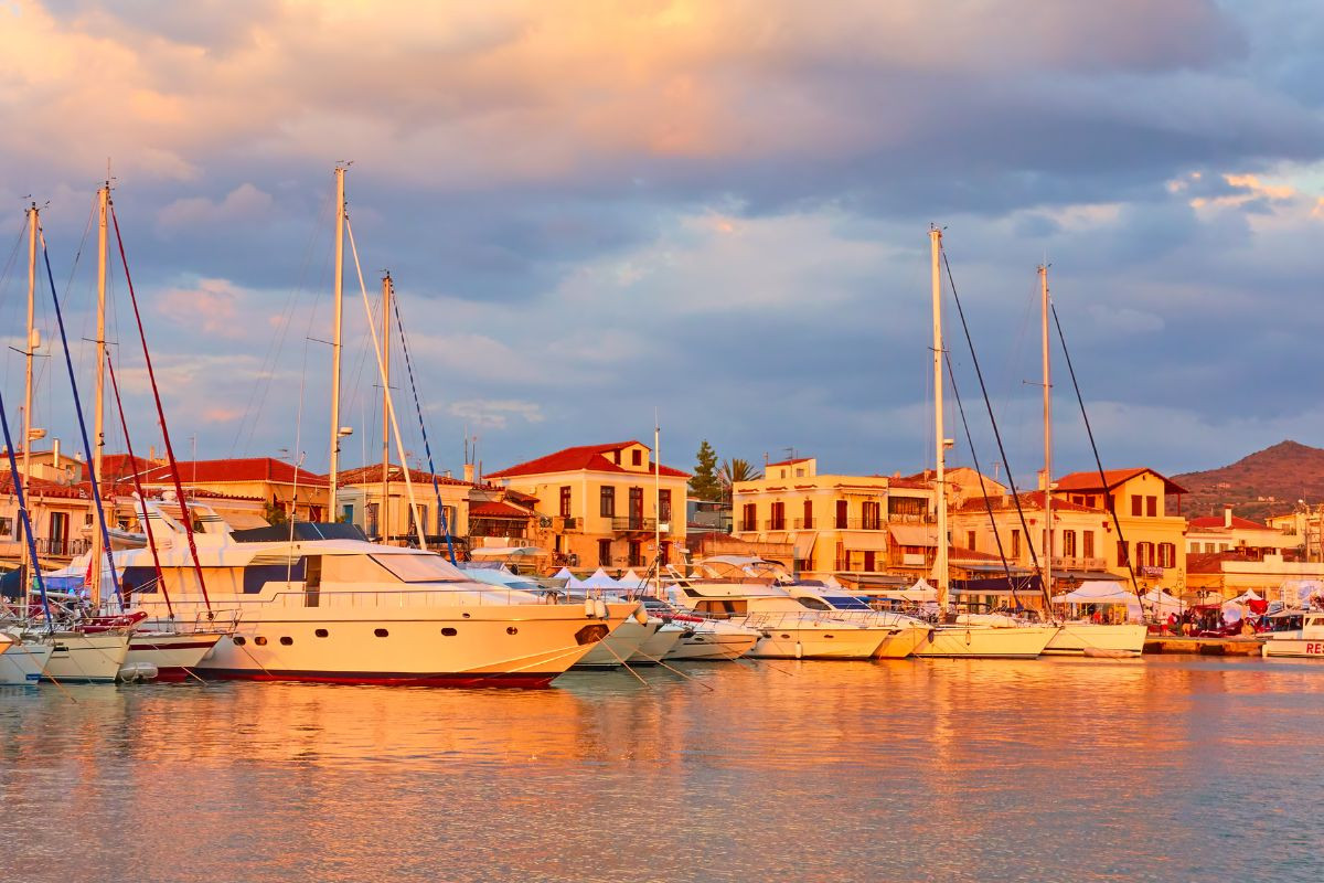 A serene view of Aegina’s harbor at sunset, with yachts docked along the waterfront and traditional buildings in the background under a colorful sky.